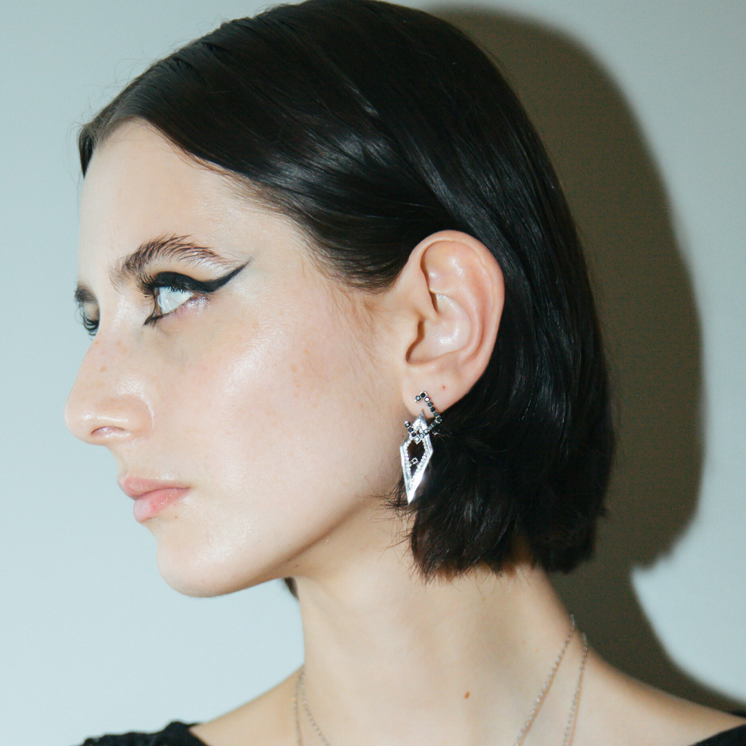 Model wearing Sadu-inspired white gold earrings with black stones and diamonds, styled with bold eyeliner and sleek short hair.