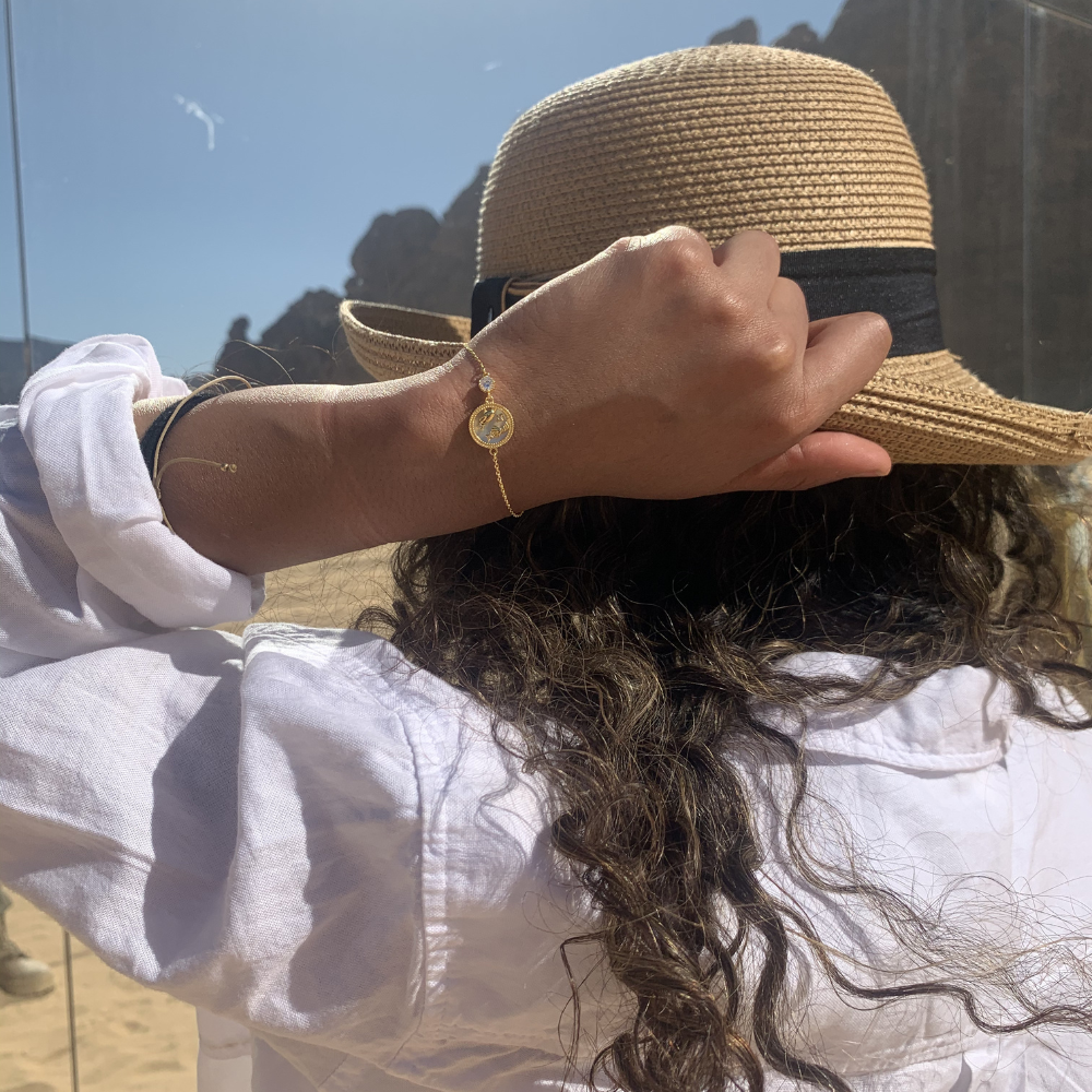 Woman wearing a gold zodiac bracelet with a wide-brim hat, arm lifted under the desert sun with scenic mountains in the background.