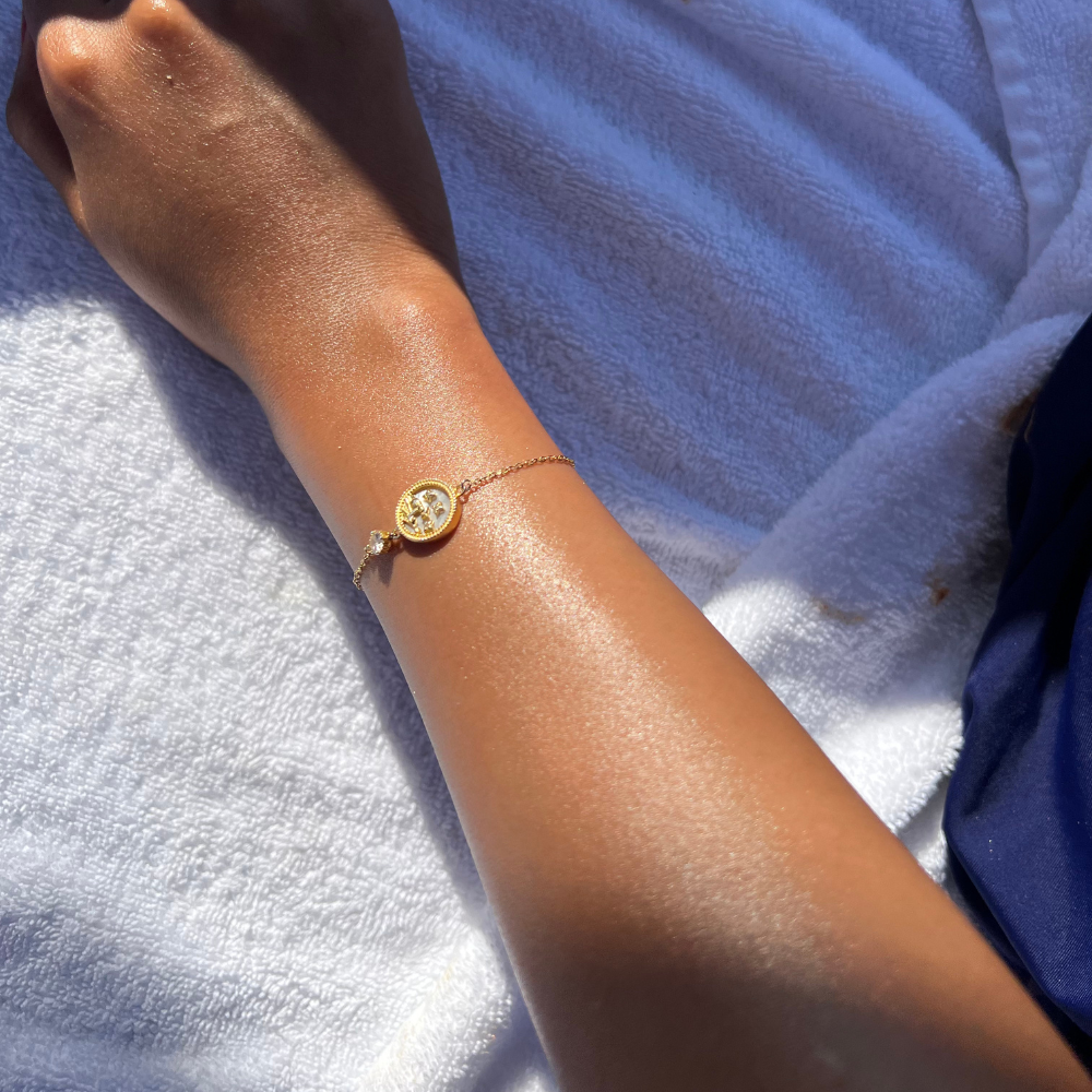 Close-up of a gold zodiac bracelet on a sun-kissed wrist, captured under natural light against a soft towel background