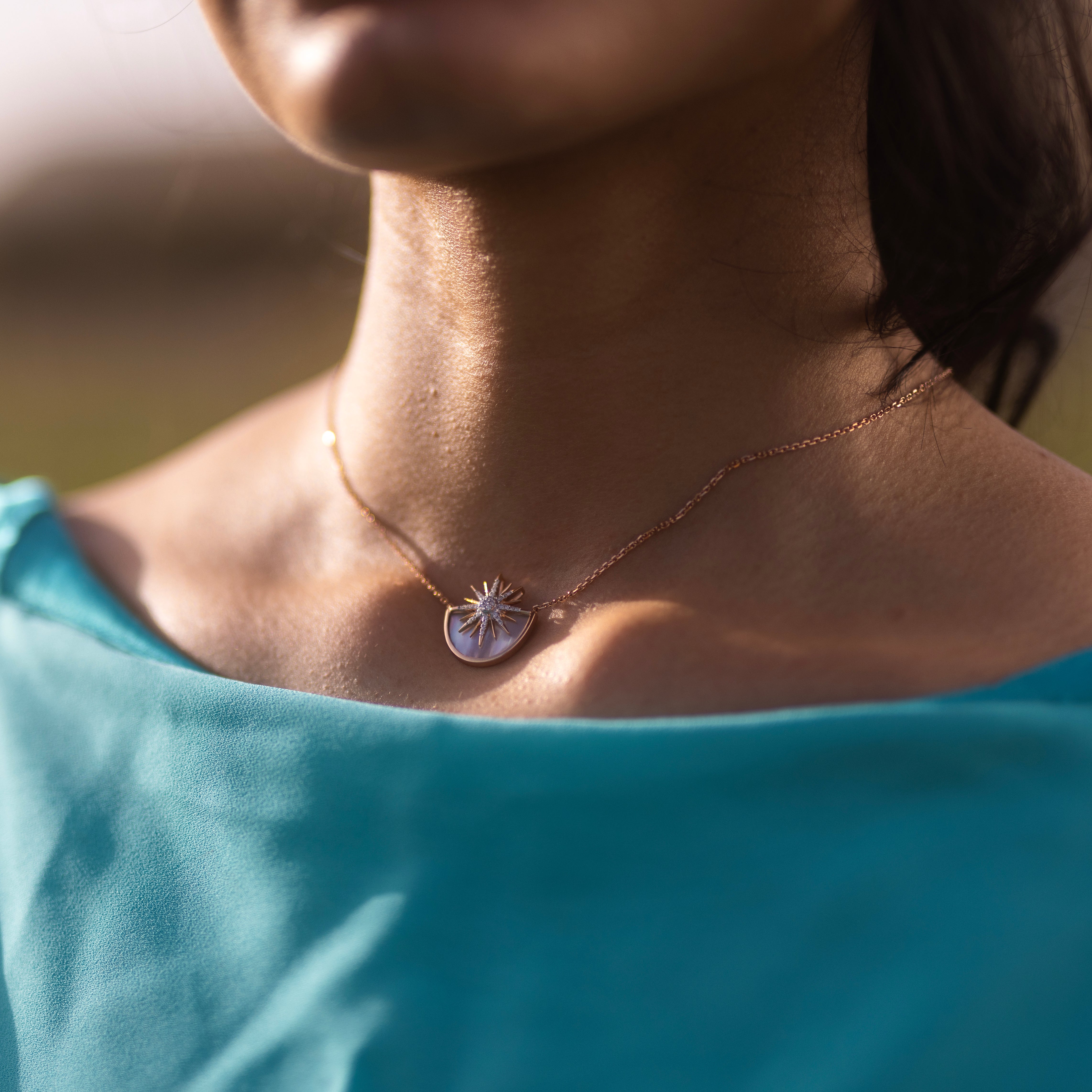 Woman wearing a rose gold necklace with a sunshine necklace made of white mother of pearl and diamonds, styled with a turquoise top.